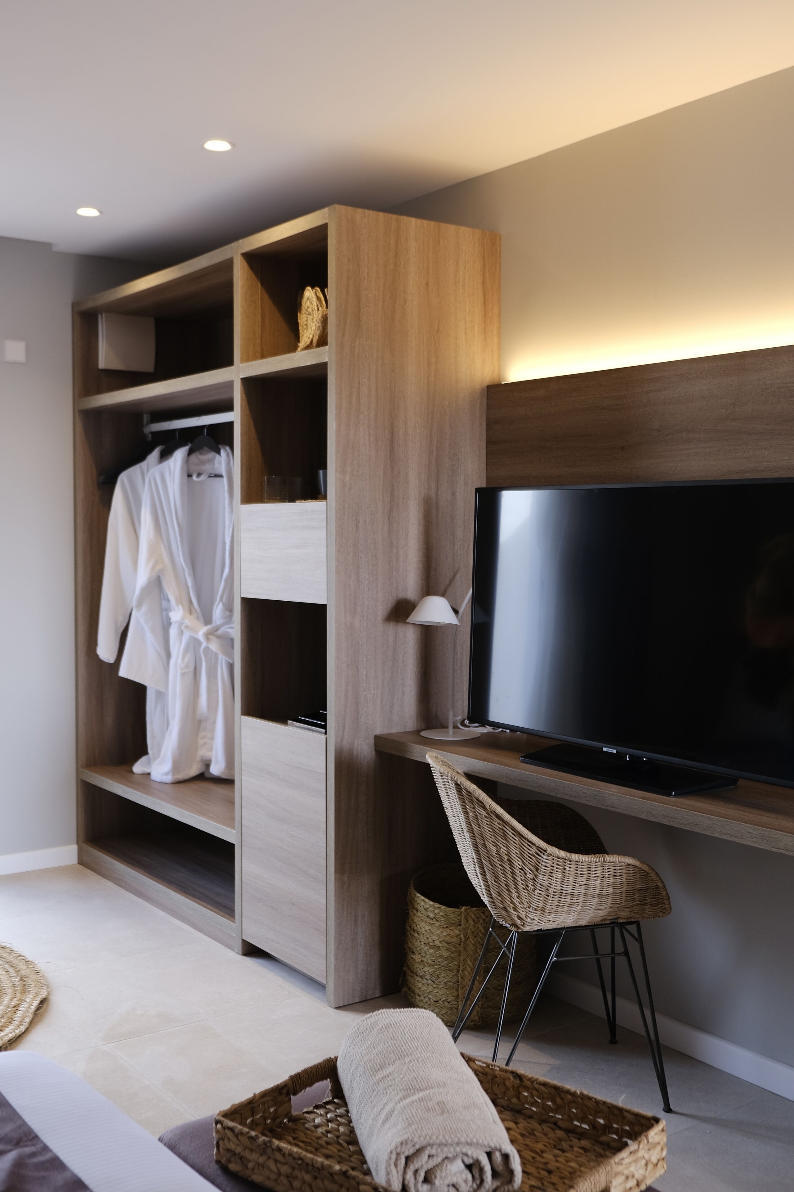 A modern room with a wooden wardrobe, a flat-screen TV, a wicker chair, and a bed with a rolled-up towel.