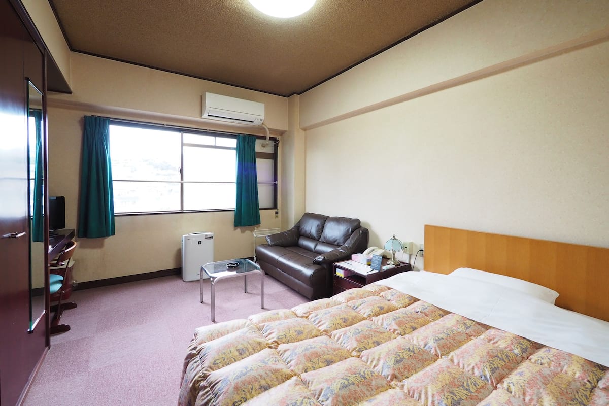 Une chambre d’hôtel comprenant un lit, un bureau, une chaise, une petite table, un radiateur et une fenêtre avec des rideaux.