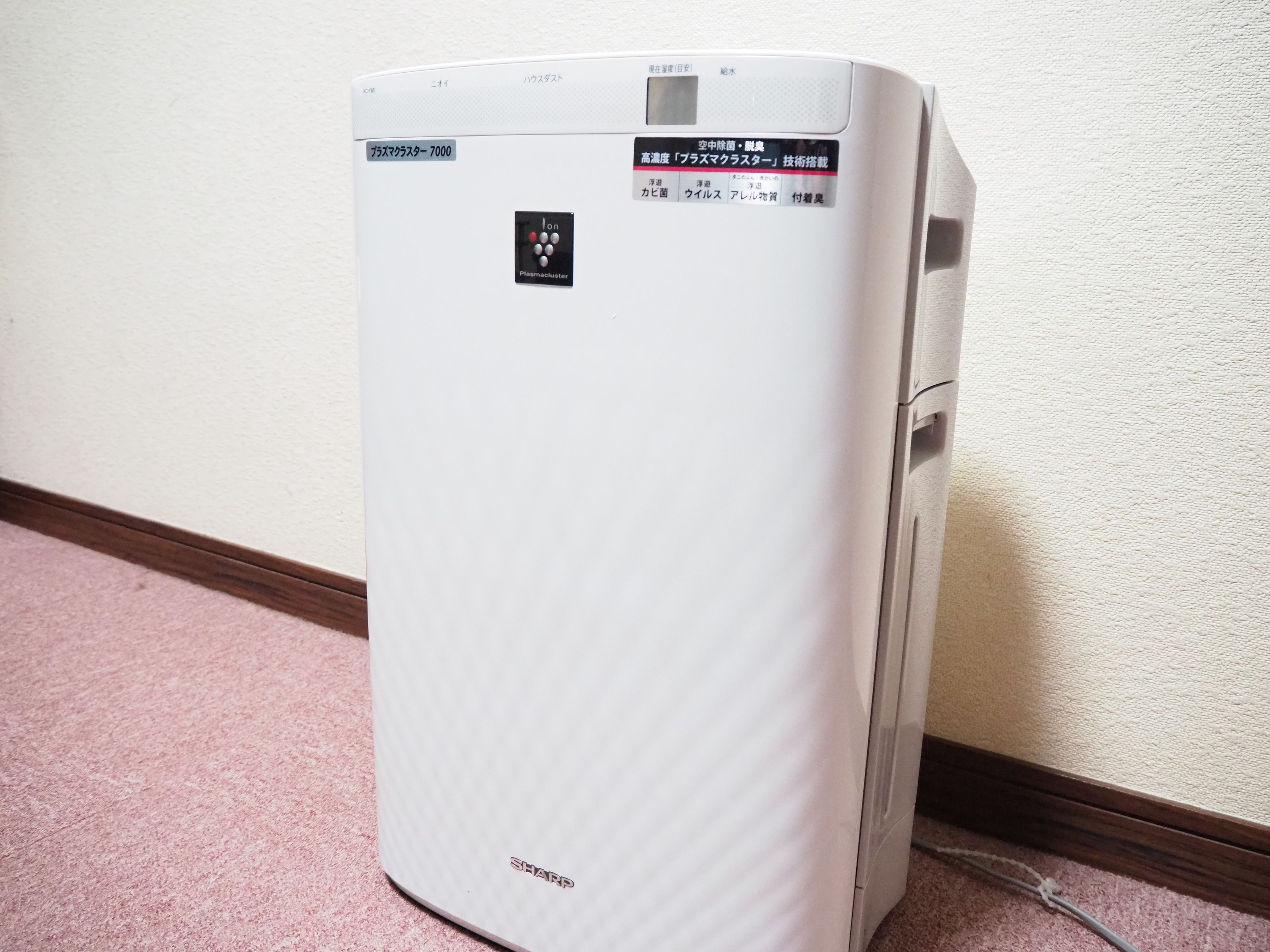 A white, modern air conditioning unit with Japanese text, placed against a wall with a wooden baseboard and a pink carpet.