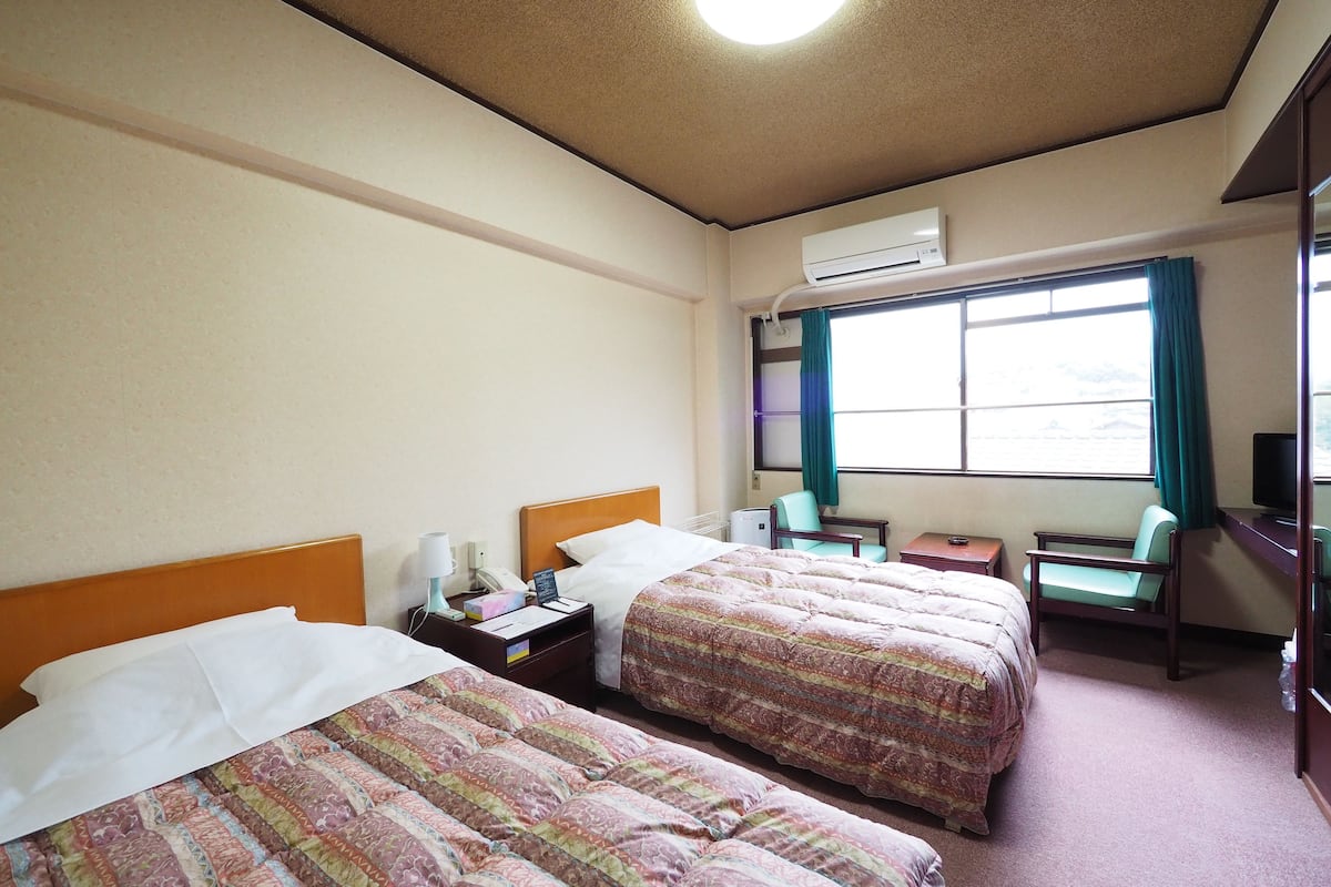 Une chambre d’hôtel avec deux lits, un bureau, une chaise, un appareil de climatisation et une fenêtre avec des rideaux.