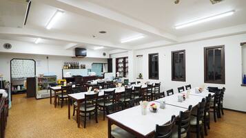 A spacious dining area with multiple tables and chairs, a kitchen area with various appliances and shelves, and a ceiling with recessed lighting.