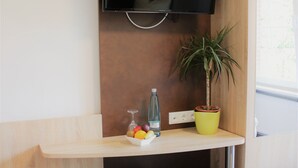 A modern room with a flat-screen TV mounted on a wooden wall, a small wooden bench, a trash bin, a potted plant, and a fruit bowl on a wooden shelf. - Hotel Restaurant Tüxen (Rendsburg)