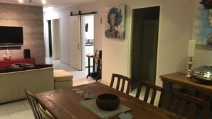 A dining area with a wooden table, chairs, and a bowl. There is a television on the wall and a door leading to another room. - Amazing apartments 2025 (Willemstad)
