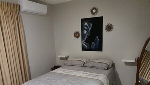 A bedroom with a bed, a nightstand, a wall-mounted air conditioner, a mirror, and decorative wall art. - Amazing apartments 2025 (Willemstad)