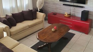 A living room with a beige sofa, a wooden coffee table, a red entertainment unit, a flat-screen TV mounted on a wooden wall, and a standing fan. - Amazing apartments 2025 (Willemstad)