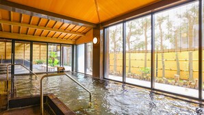 Zona de baño de aguas termales con vigas de madera, una escalera metálica y ventanas amplias con vistas a una valla de bambú.