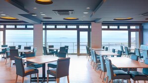 Un comedor amplio con mesas y sillas, ventanas grandes que ofrecen vista al mar, y un diseño moderno en el techo.