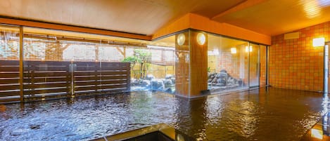Un onsen japonais traditionnel, avec un intérieur en bois, une source d’eau chaude entourée de pierres et une vue sur un paysage naturel grâce à une paroi en verre.