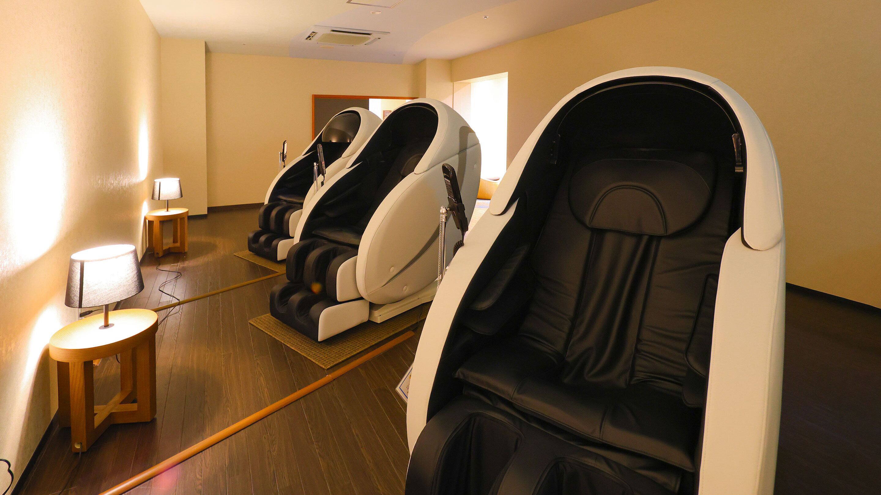 A room with massage chairs, a small table with a lamp, and wooden flooring.