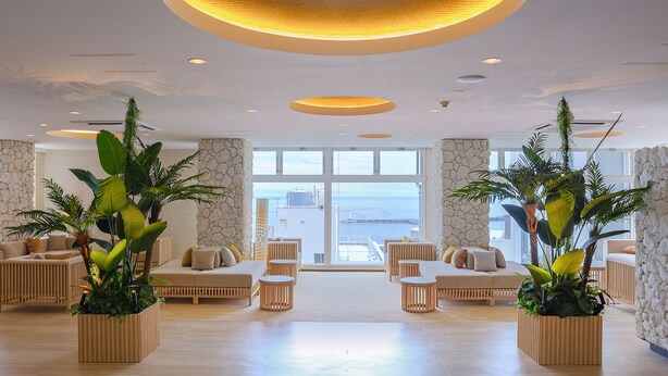 Un vestíbulo espacioso con techo alto, amplias ventanas que ofrecen vistas al mar y varias áreas de descanso con plantas en macetas.