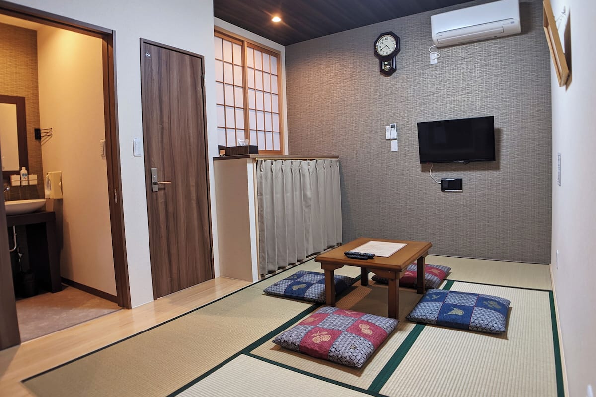 A traditional Japanese room with tatami flooring, a wooden table, cushions, a television, and a clock on the wall.