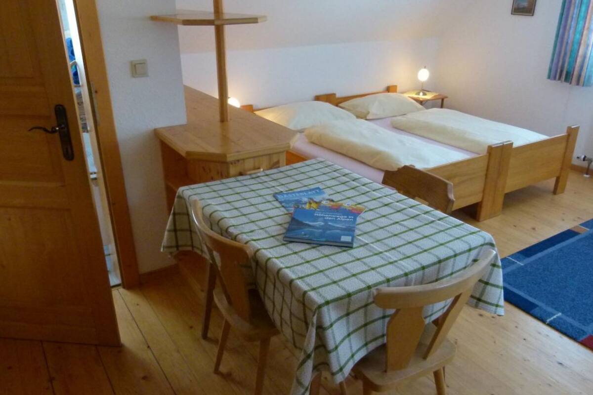 Una habitación pequeña con una mesa de madera y sillas, una cama con colchón y almohadas blancas, y una mesita de noche con lámpara.