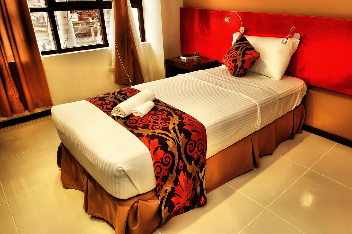 Cama bem arrumada com edredom branco e manta decorativa estampada, cabeceira vermelha e mesinha de cabeceira com um livro e uma luminária.