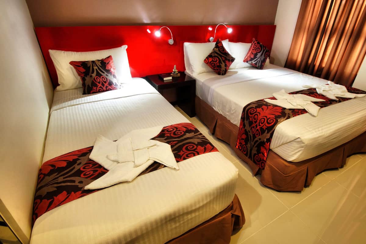 Quarto de hotel com duas camas, cada uma com colcha branca e mantas decorativas estampadas com flores vermelhas. Cortinas e cabeceira vermelhas.