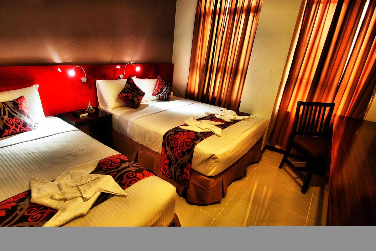 Quarto de hotel com duas camas de solteiro, uma mesinha de cabeceira, uma cadeira e uma parede de destaque vermelha.