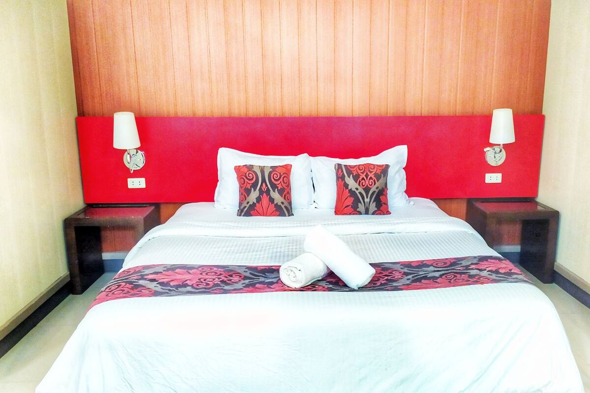 Quarto de hotel com cabeceira vermelha, duas mesas de cabeceira com luminárias, uma cama grande com colcha florida e dois travesseiros decorativos.