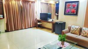 Ein Wohnzimmer mit einer Couch, einem Glastisch mit Pflanze, einem Fernseher, einem kleinen Kühlschrank und einem Schreibtisch mit Stuhl.