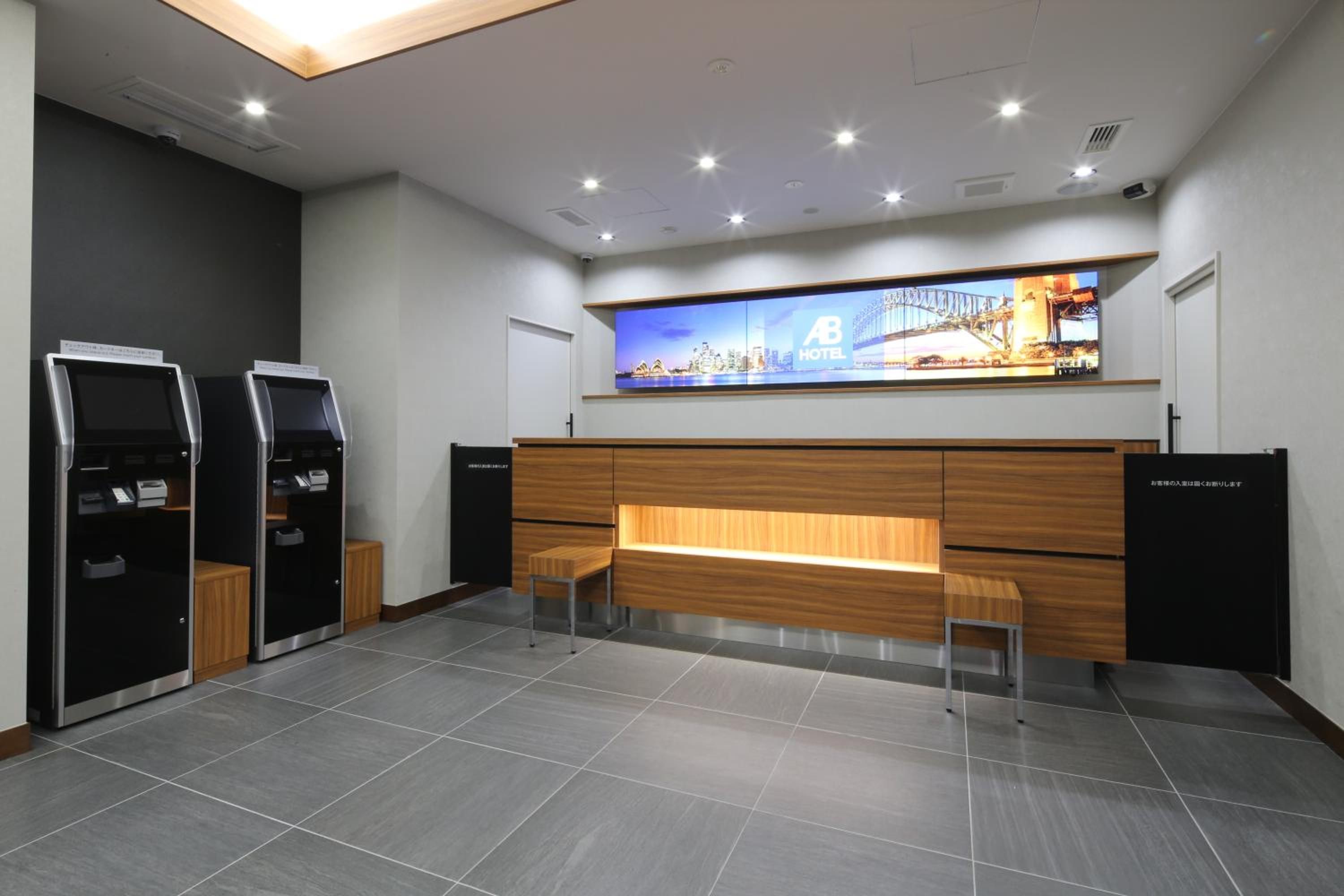 A hotel reception area with a modern design, featuring a reception desk, two self-service kiosks, and a large digital display above the desk.