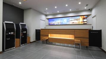 A hotel reception area with a modern design, featuring a reception desk, two self-service kiosks, and a large digital display above the desk.