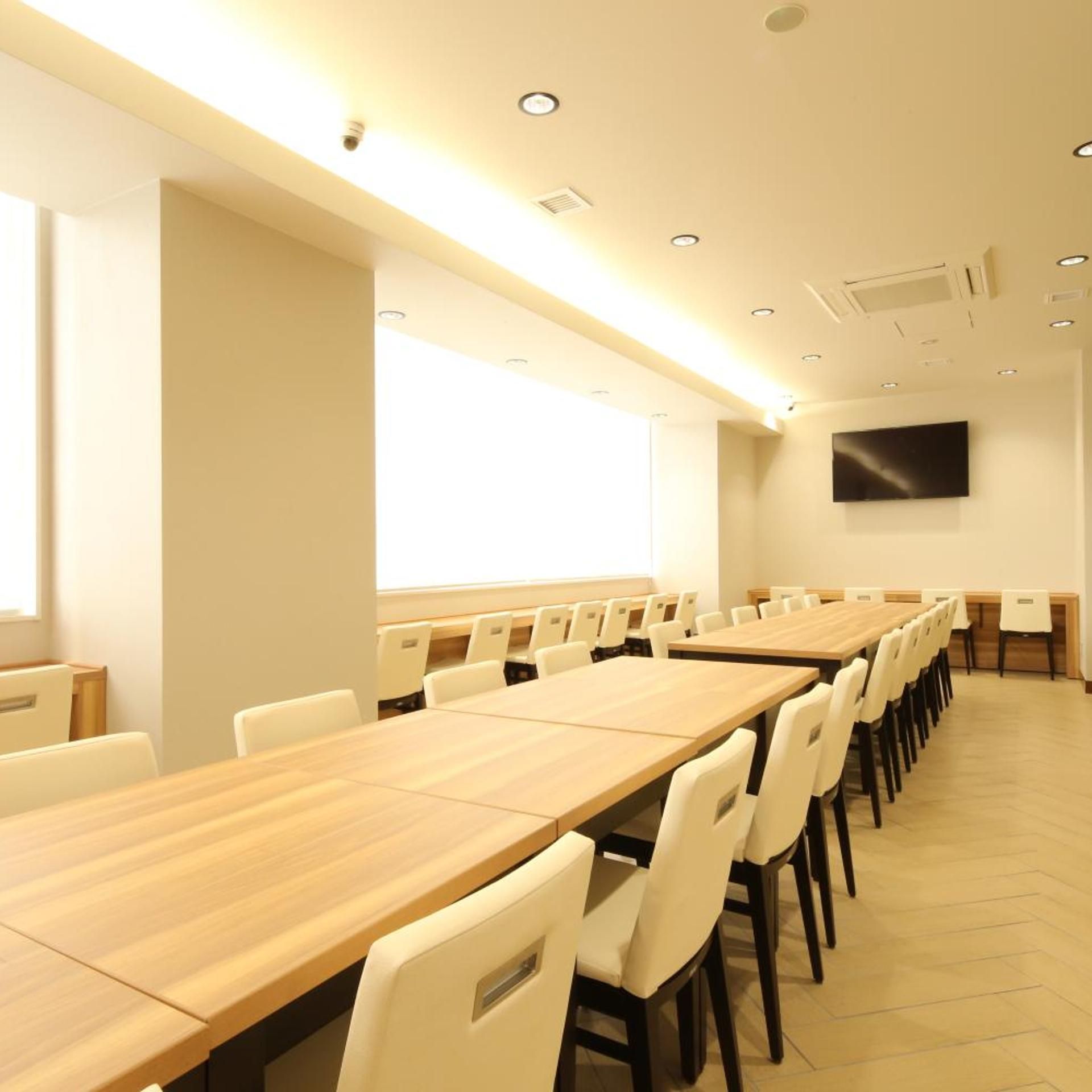 A conference room with long tables and chairs, a flat-screen TV mounted on the wall, and a window providing natural light.