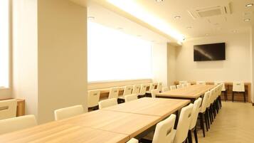 A conference room with long tables and chairs, a flat-screen TV mounted on the wall, and a window providing natural light.