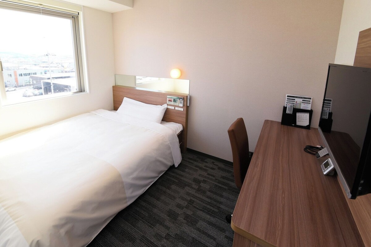 Ett hotellrum med en säng, ett skrivbord, en stol, en skrivbordslampa och utsikt över staden genom fönstret.