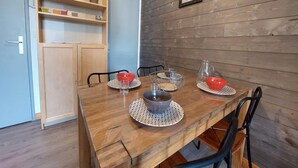 Ein Essbereich mit einem Holztisch, gedeckt für vier Personen, umgeben von Stühlen. Links befindet sich eine Holzkommode mit Regalen, und an der Wand hängen zwei gerahmte Bilder.