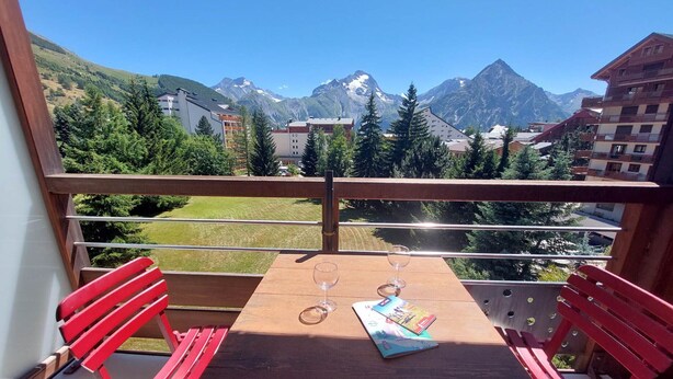 Ein Balkon mit Holztisch und roten Stühlen, mit Blick auf eine gebirgige Landschaft und Gebäude im Hintergrund.