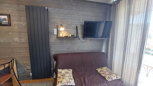 Ein Zimmer mit einem braunen Sofa, zwei gemusterten Kissen, einem angebrachten Fernseher und einem kleinen Tisch mit einer Lampe.
