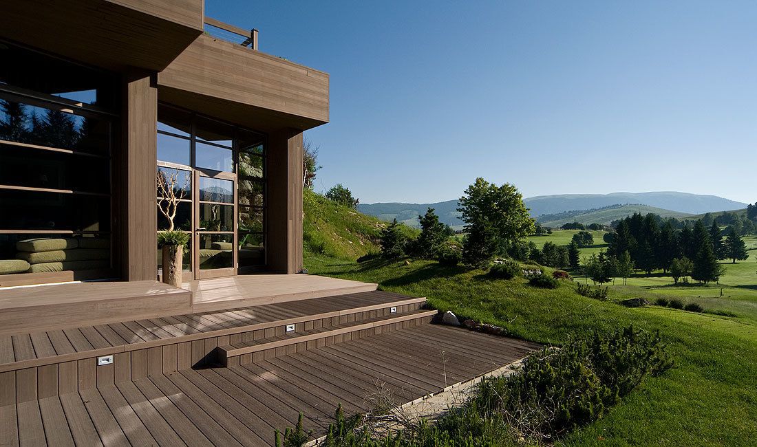 A modern house with a wooden deck and large windows overlooking a green landscape.