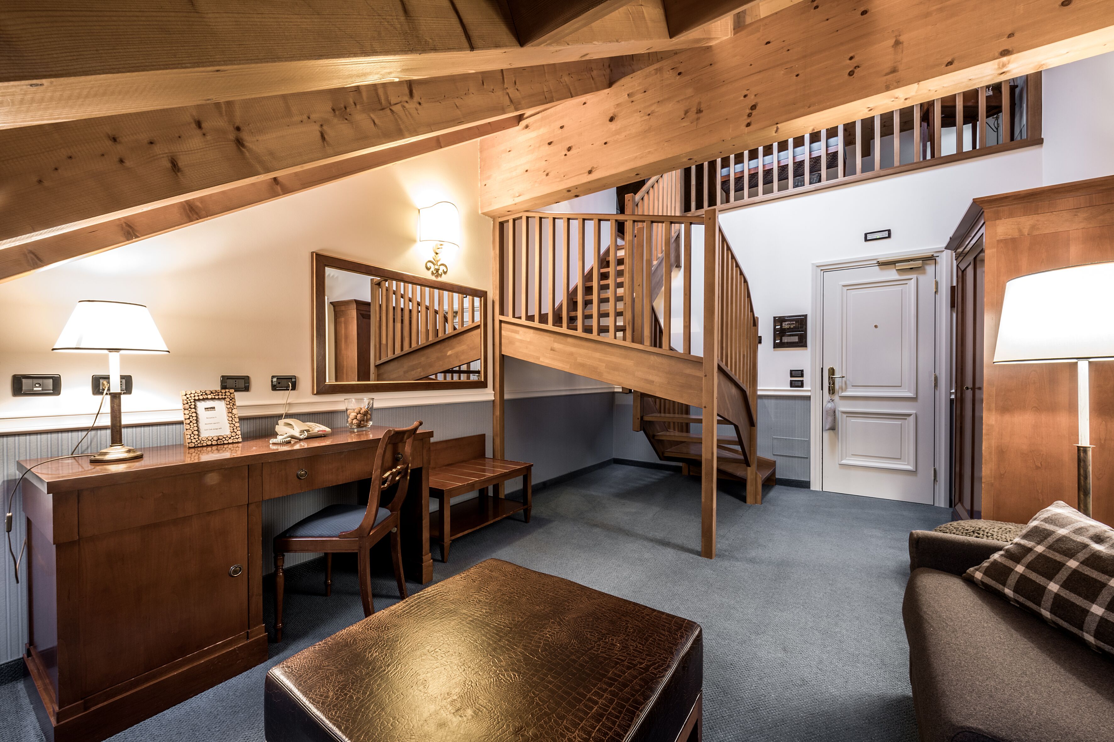 A cozy attic bedroom with a wooden desk, chair, and a small table. A wooden staircase leads to an upper level.