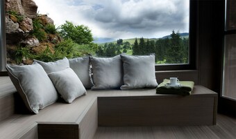 Una habitación moderna con una zona de estar con bancos integrados, una ventana con vistas a un bosque y colinas, y una mesita con una taza y una toalla verde doblada.