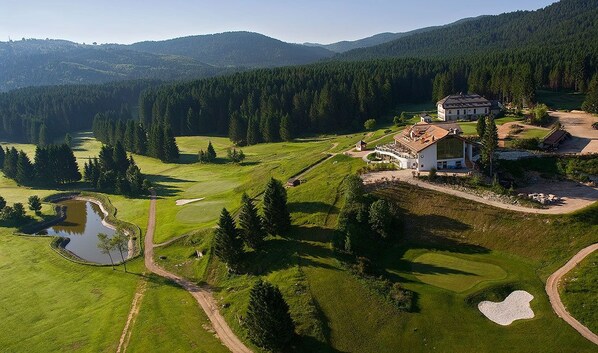 An aerial view of a golf course with a clubhouse, a pond, and surrounding forested hills. - Meltar Boutique Hotel (Asiago)