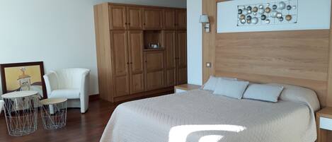 Un dormitorio con una cama grande, un armario de madera, un sillón blanco, una mesita redonda y una obra de arte en la pared.
