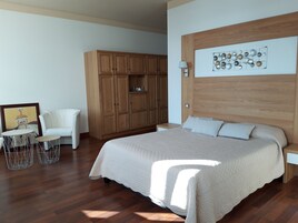 Un dormitorio con una cama grande, un armario de madera, un sillón blanco, una mesita redonda y una obra de arte en la pared.
