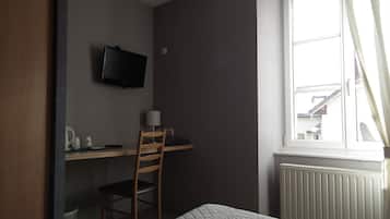 Una habitación pequeña y tenuemente iluminada, con una cama, un escritorio, una silla y un televisor montado en la pared.