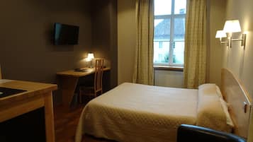 A hotel room with a bed, a desk with a chair, a television on the wall, and a window with curtains.
