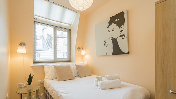 A neatly made bed with white linens, a bedside table with a lamp, and a window with blinds. - Rose Street Apartment City Centre (Edinburgh)