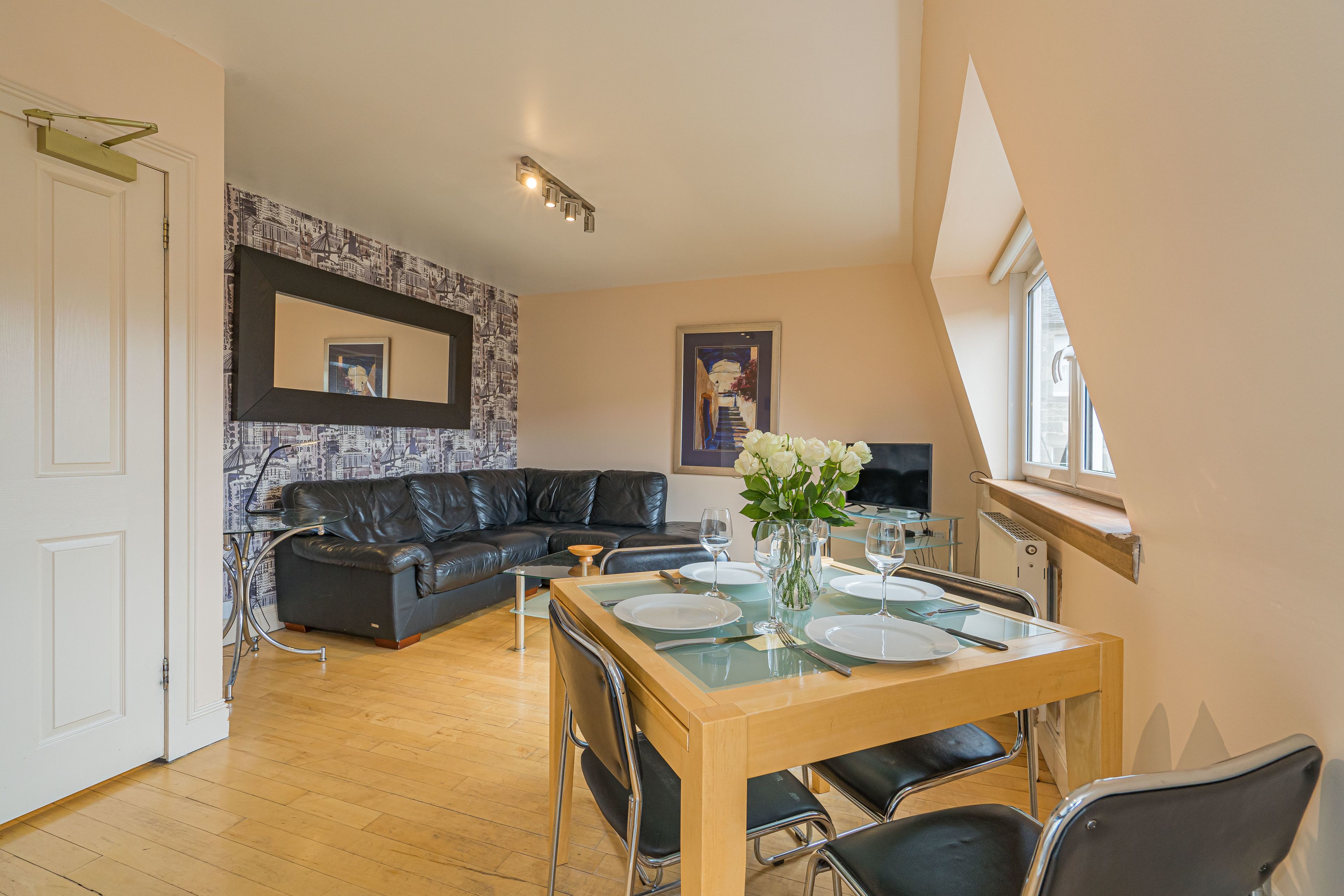 A modern living room with a wooden dining table set for four, a black leather sofa, and a cityscape wallpaper.