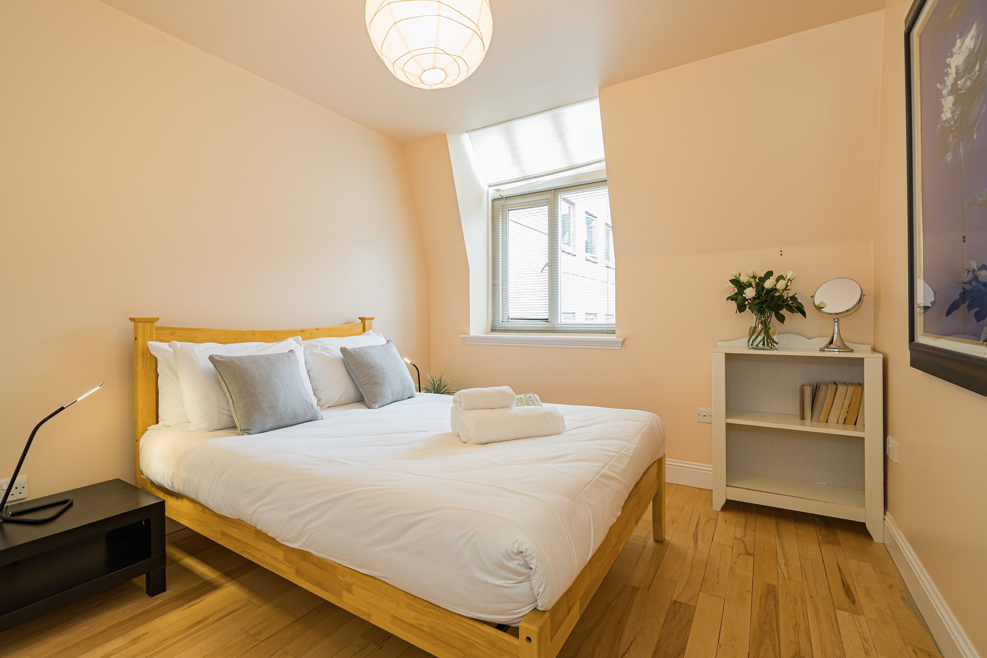 Un lit bien fait, avec du linge de lit blanc et des oreillers, une table de chevet avec une lampe, une petite étagère avec des livres et une plante, et une fenêtre avec des stores.