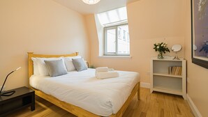 Ein ordentlich bezogenes Bett mit weißen Leinen und Kissen, ein Nachttisch mit Lampe, ein kleines Regal mit Büchern und einer Pflanze sowie ein Fenster mit Jalousien.