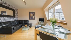 Ein modernes Wohnzimmer mit einem schwarzen Ledersofa, einem Esstisch für zwei Personen und einem großen Fenster mit Blick auf Gebäude.