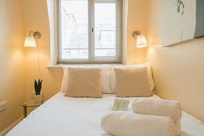 A hotel room with a bed, two bedside lamps, a window with blinds, a small table with a plant, and a wall-mounted artwork.