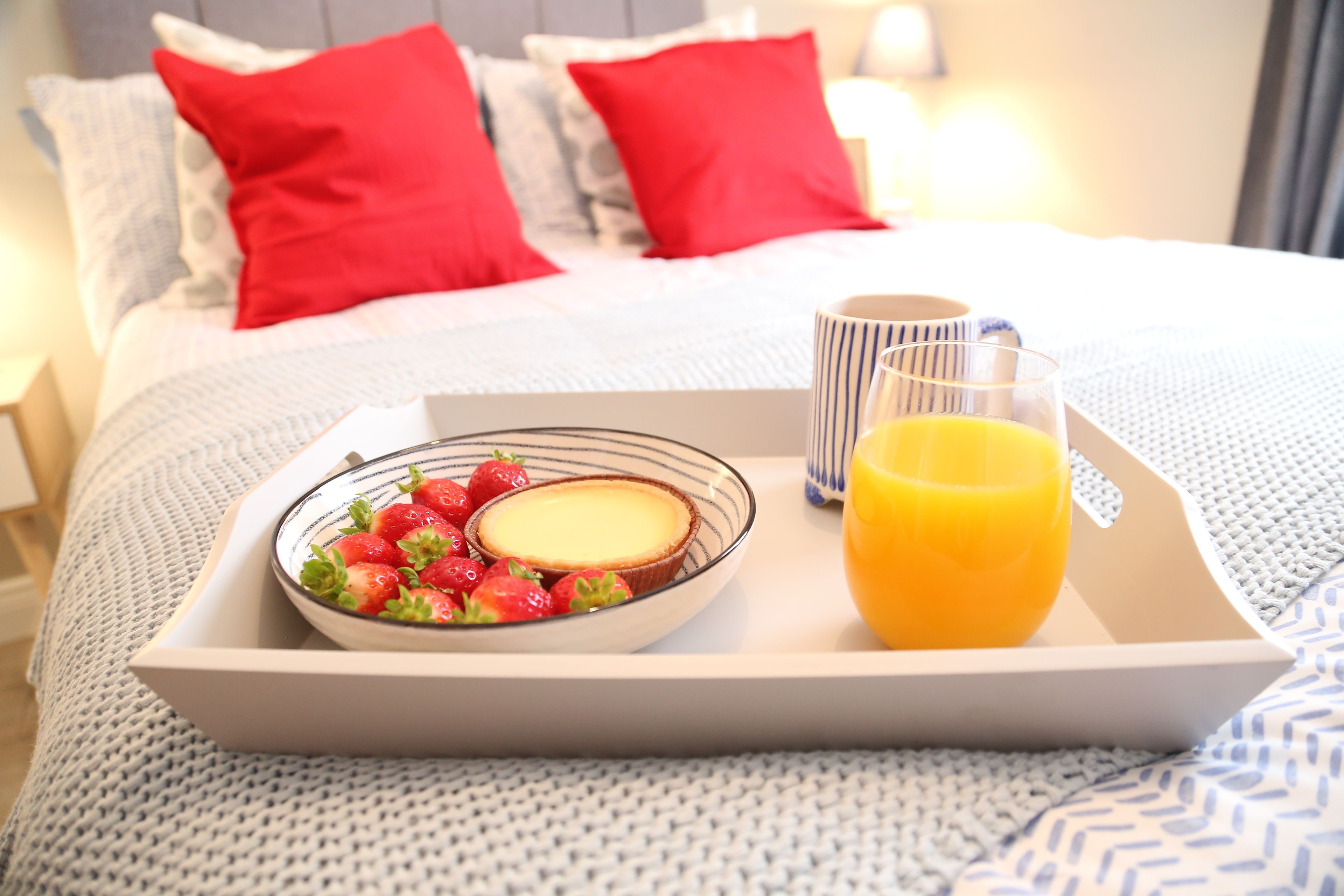 A breakfast tray with strawberries, a slice of cheesecake, and a glass of orange juice on a bed with red pillows.