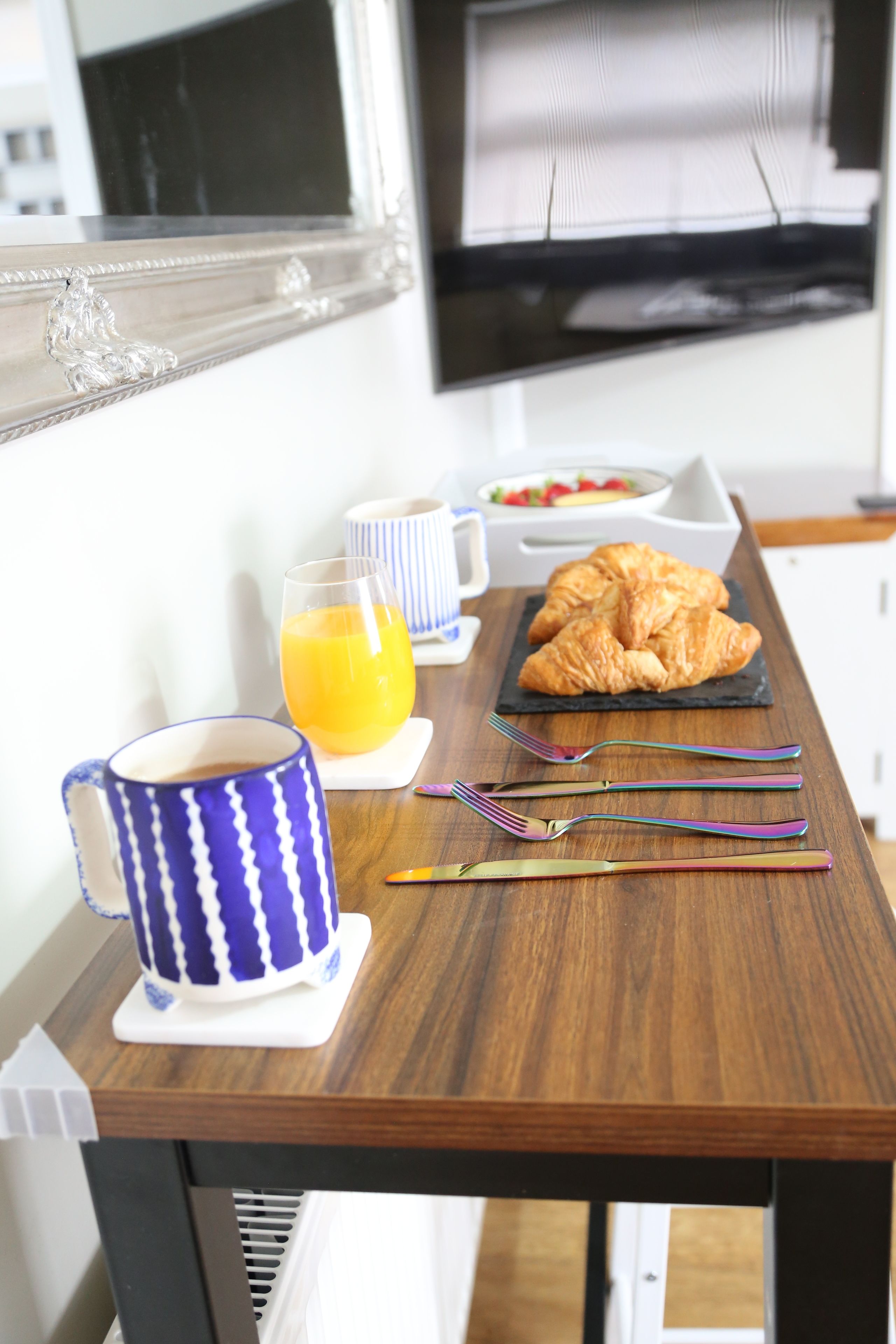 A breakfast table with a croissant, a glass of orange juice, a blue and white mug, and a tray with utensils.