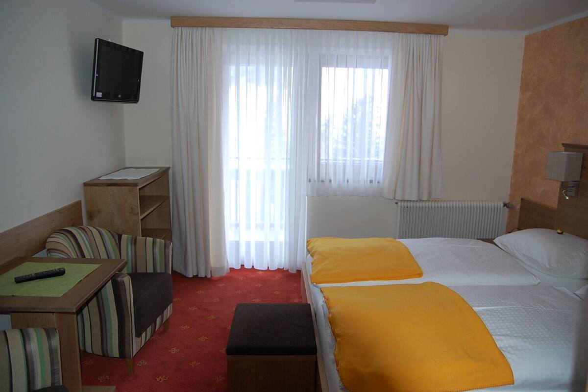 A hotel room with a bed, a desk, a chair, a small table, a TV, and a window with curtains.