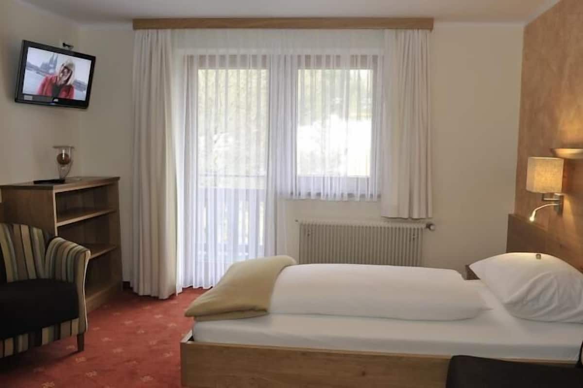 A hotel room with a single bed, a chair, a small table, a TV on the wall, and a window with sheer curtains.
