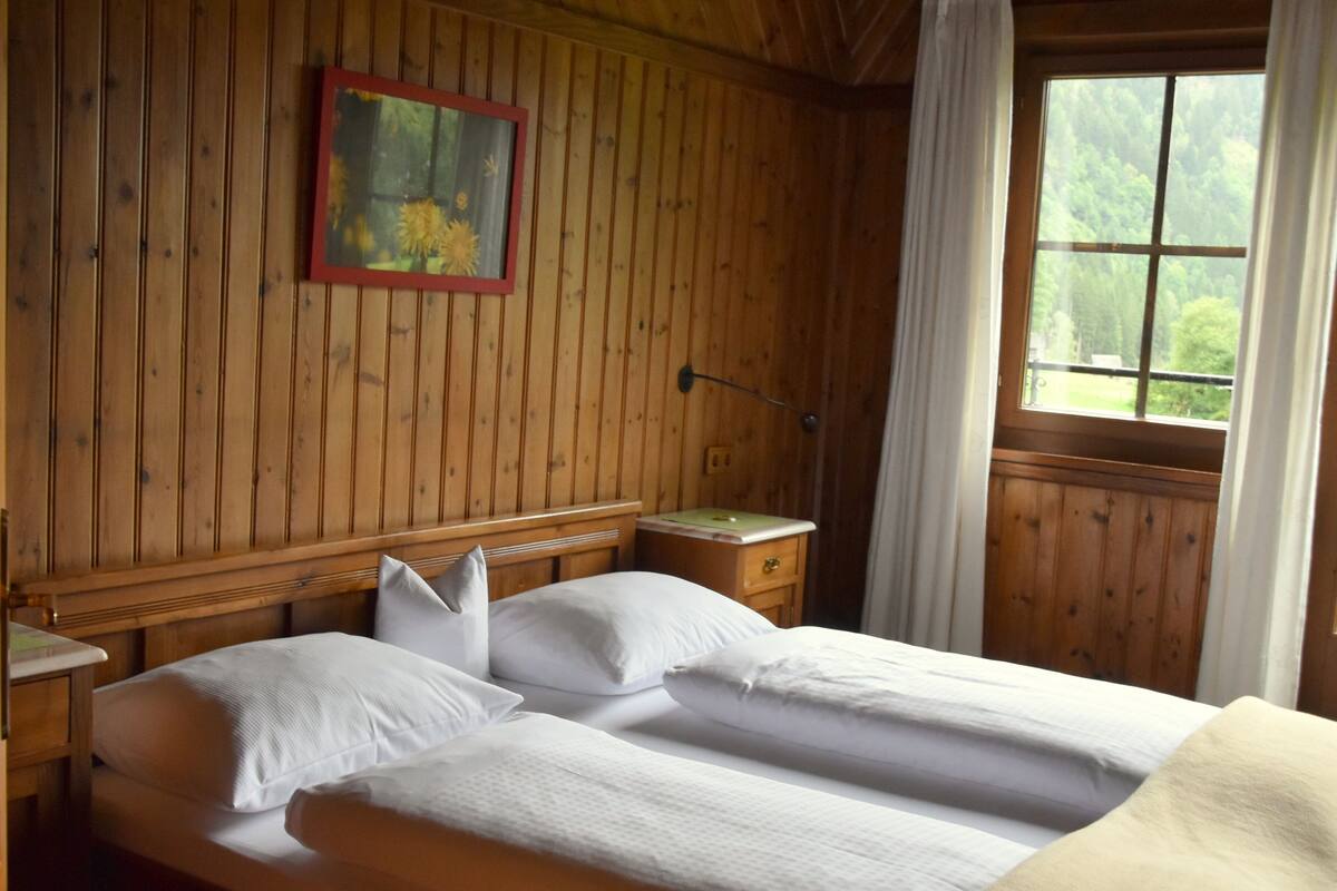 A wooden room with a bed, a nightstand, a lamp, and a window with curtains.