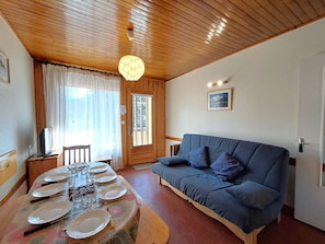 A room with a dining table set for four, a blue sofa, wooden ceiling, and a door leading outside. - Apartment 8 guests, near slopes, pets allowed (Les Deux Alpes)
