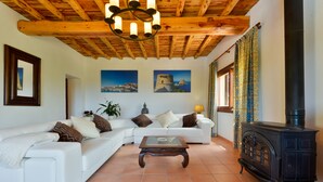 Ein Wohnzimmer mit einem weißen Sofa, einem Glastisch und einer Holzbalkendecke. Rechts befindet sich ein schwarzer Herd und ein Gemälde an der Wand.